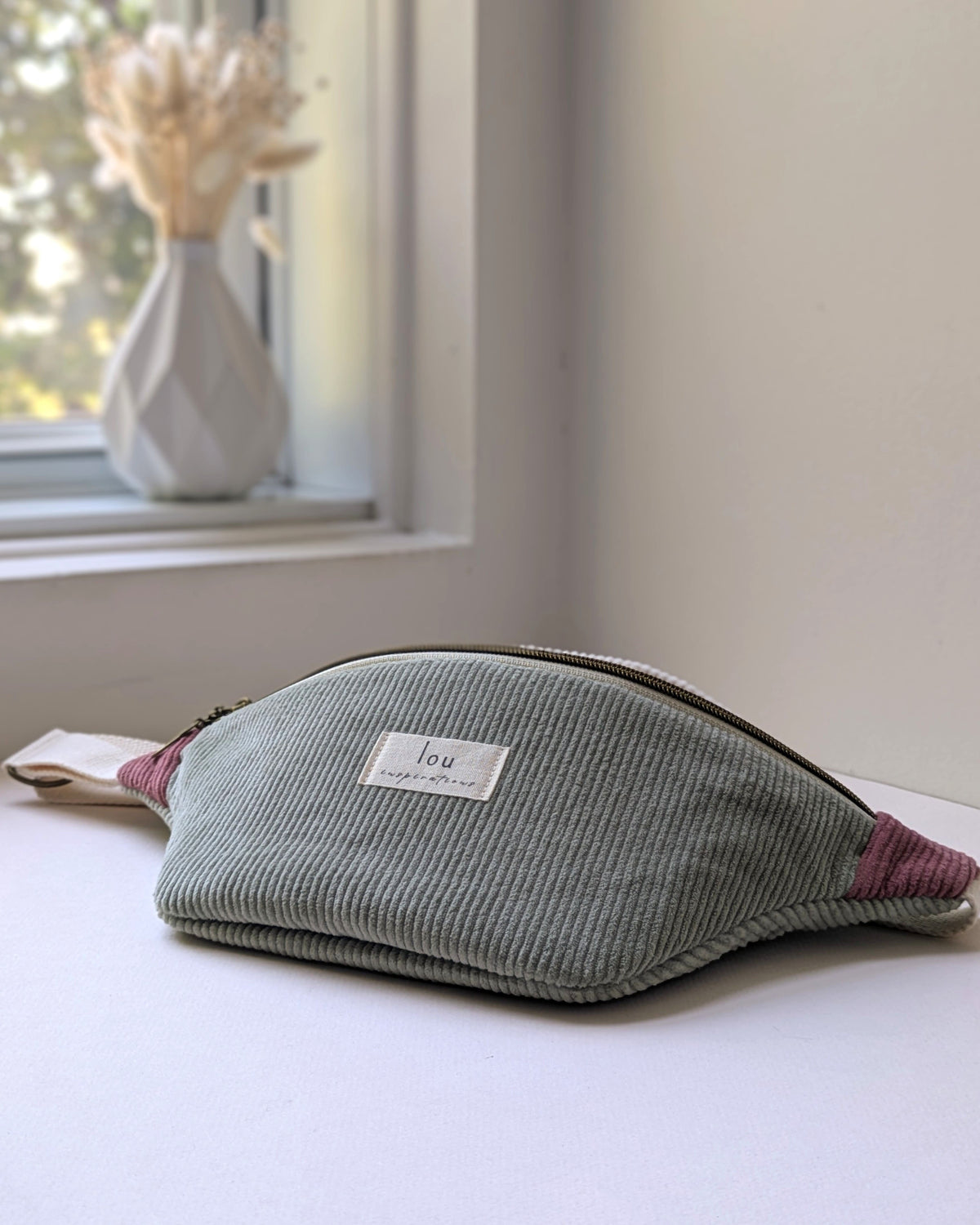 Sac banane tricolore en velours côtelé vert sauge avec accents vieux rose et étiquette Lou Inspirations, posé près d’un vase de fleurs séchées