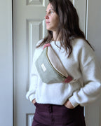 Femme portant un sac banane en velours côtelé vert sauge et vieux rose en bandoulière avec un pull blanc