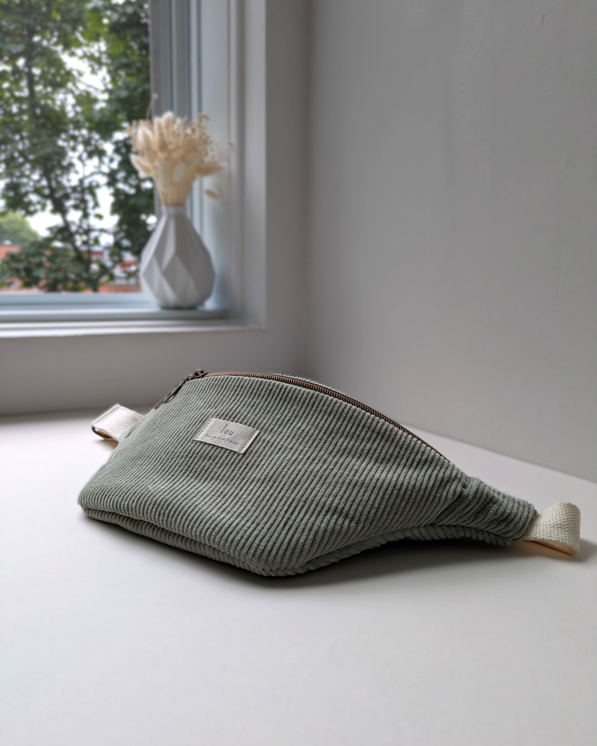 Sac banane en velours côtelé bio sauge près d’un vase de fleurs séchées avec la lumière naturelle de la fenêtre